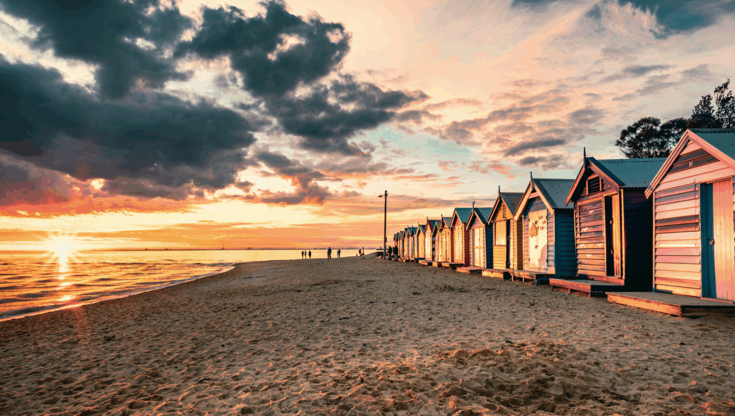 Sunset over a sandy beach with a row of colourful beach huts and holiday homes along the shoreline.