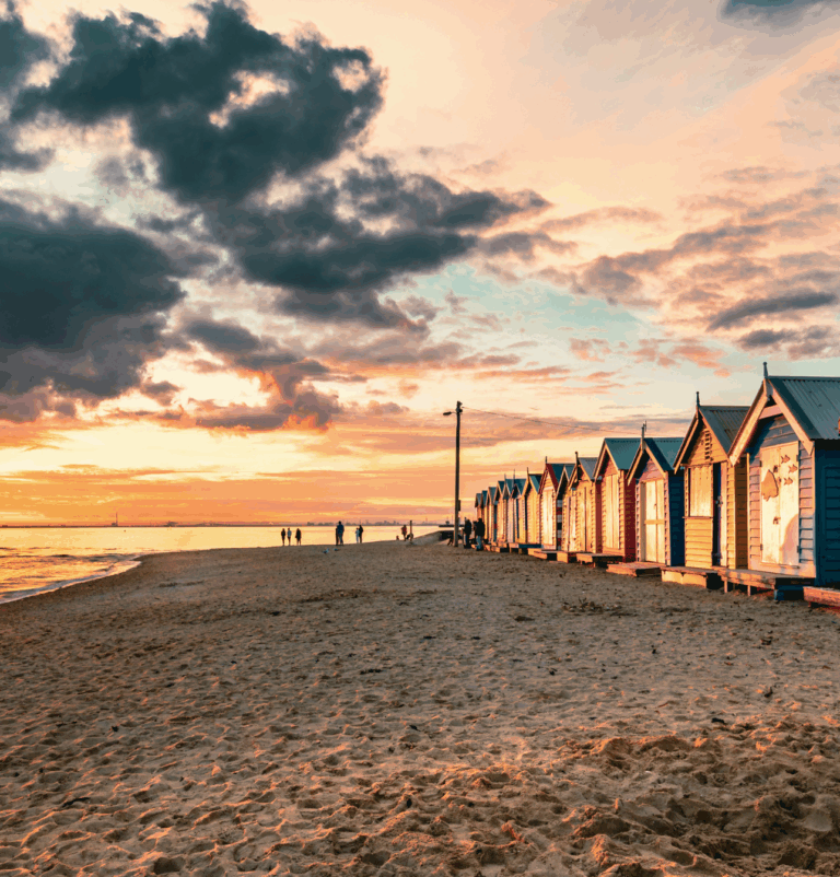 Sunset over a sandy beach with a row of colourful beach huts and holiday homes along the shoreline.